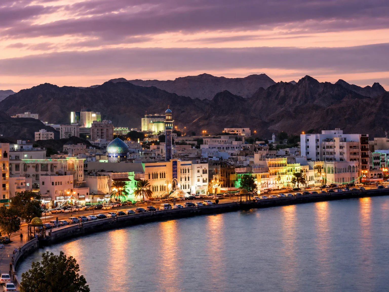 واجهة مطرح البحرية مضاءة عند الغروب وخلفها جبال مسقط الداكنة