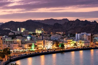 واجهة مطرح البحرية مضاءة عند الغروب وخلفها جبال مسقط الداكنة
