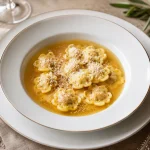 Square photograph of small round anolini floating in clear golden broth with grated Parmigiano and a restrained herb garnish.