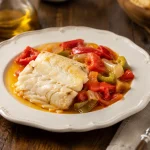 Square photograph of a white cod fillet surrounded by glossy red and green pepper strips and onion on a shallow dark plate.