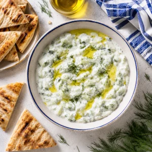 蓝边碗中的白色 Tzatziki 酸奶黄瓜酱，周围摆有烤扁面包、莳萝和橄榄油